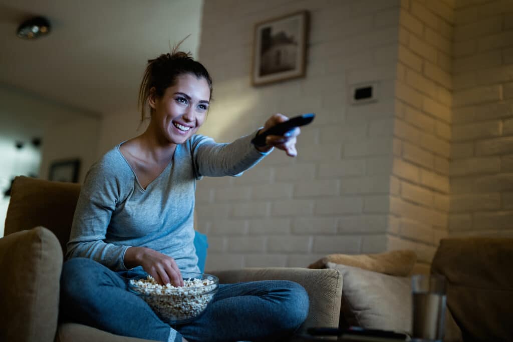 wanita muda bersenang-senang sambil mengganti saluran tv makan popcorn malam hari di rumah