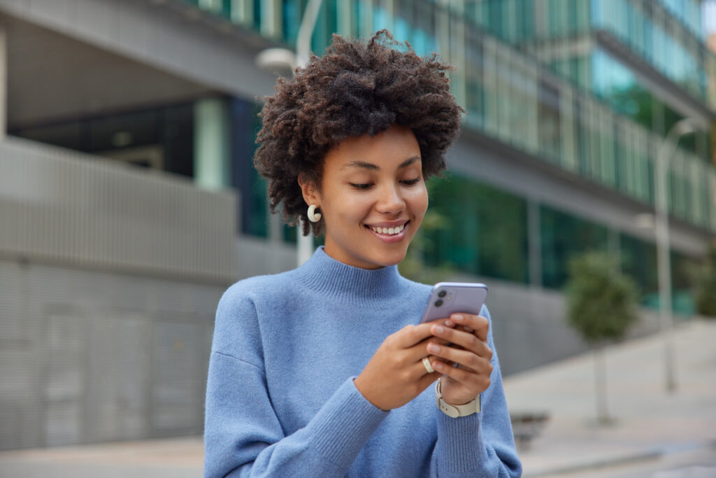 glad young woman with curly bushy hair holds modern mobile phone downloads amazing application surfs social networks wears casual blue jumper 2